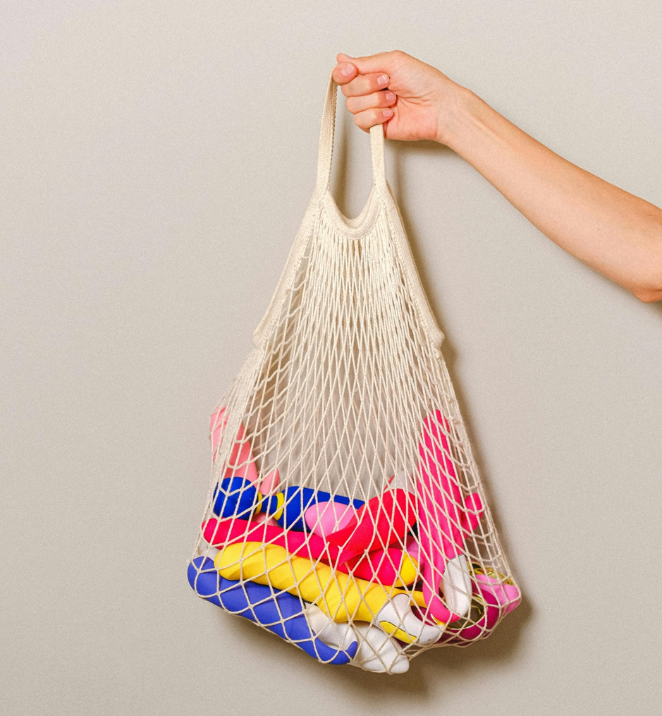 a hand holding a string bag against a beige backdrop, containing multiple vibrator sex toys in various colours